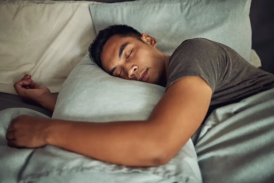 Young man sleeping peacefully on his side in bed, hugging a pillow, wearing a gray t-shirt with soft natural lighting.