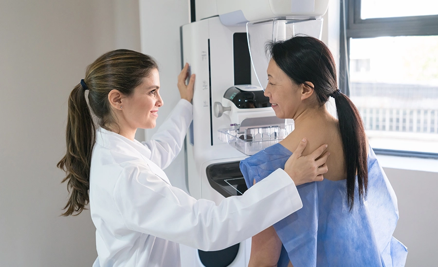 Smiling clinician in a white coat guides a woman in a blue gown into position at a mammography machine.